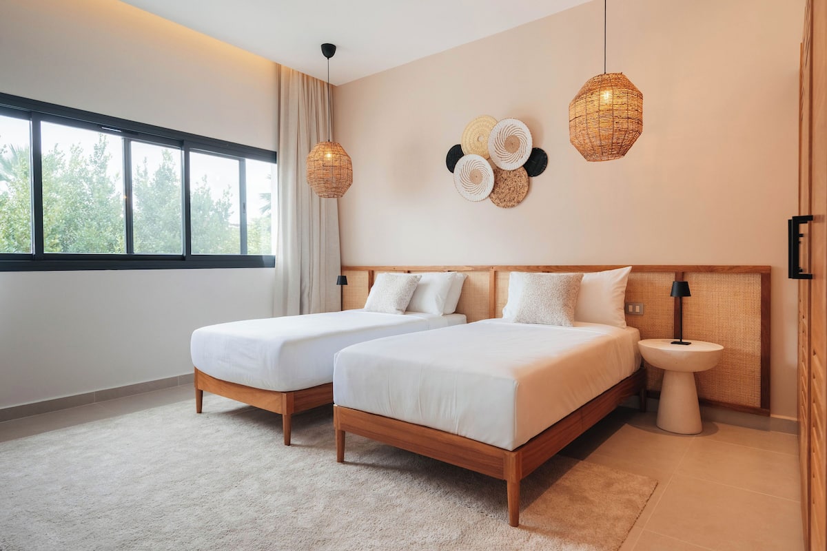 A bedroom with two beds, a bedside table, a hanging light fixture, and a window with curtains.