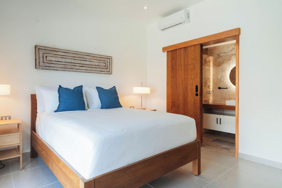 A bedroom with a wooden bed, a nightstand, a wall-mounted lamp, a wardrobe with a mirror, and a built-in air conditioning unit.