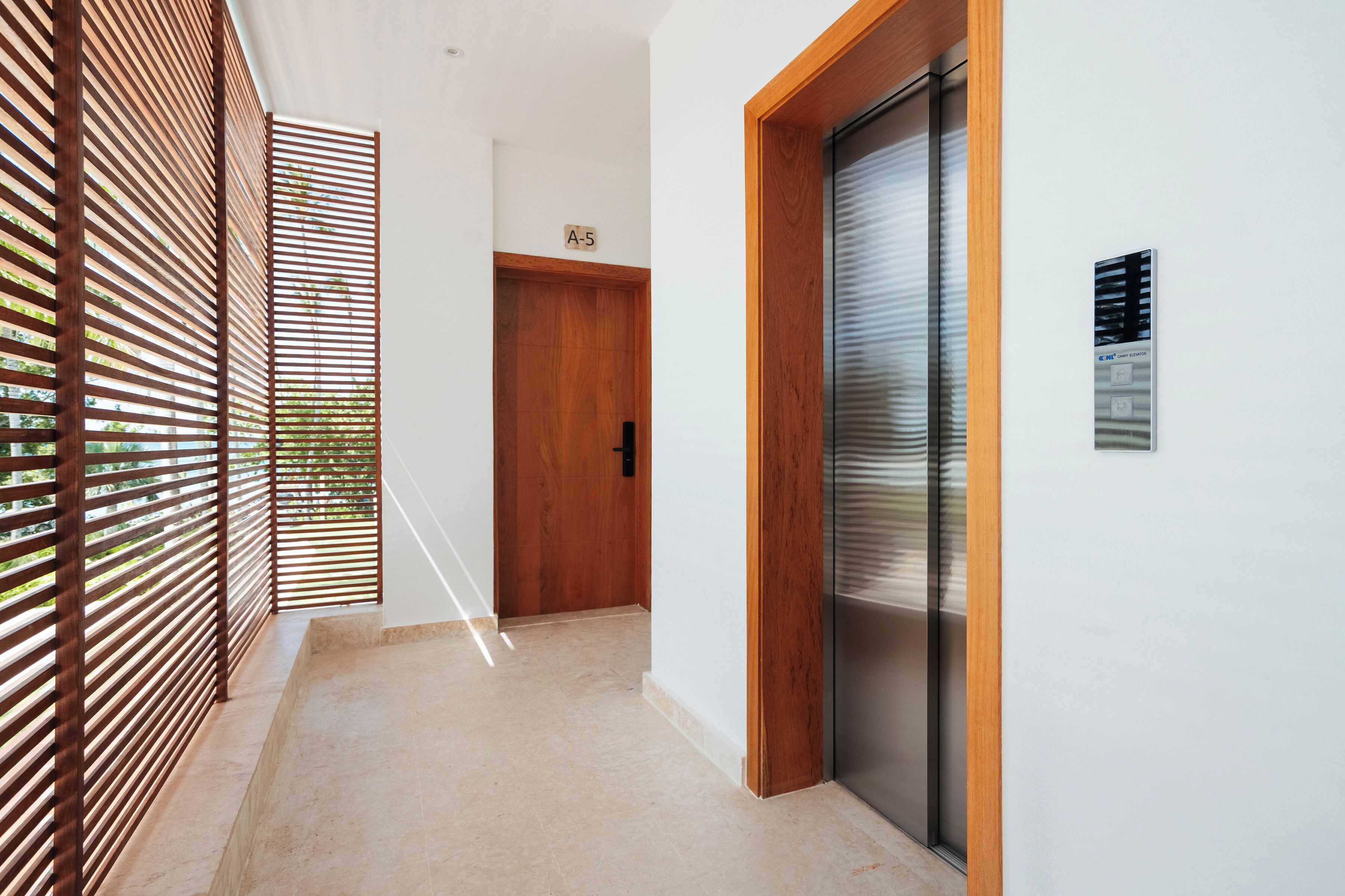 A modern hallway with a wooden door, a glass elevator, and a wooden slatted partition.