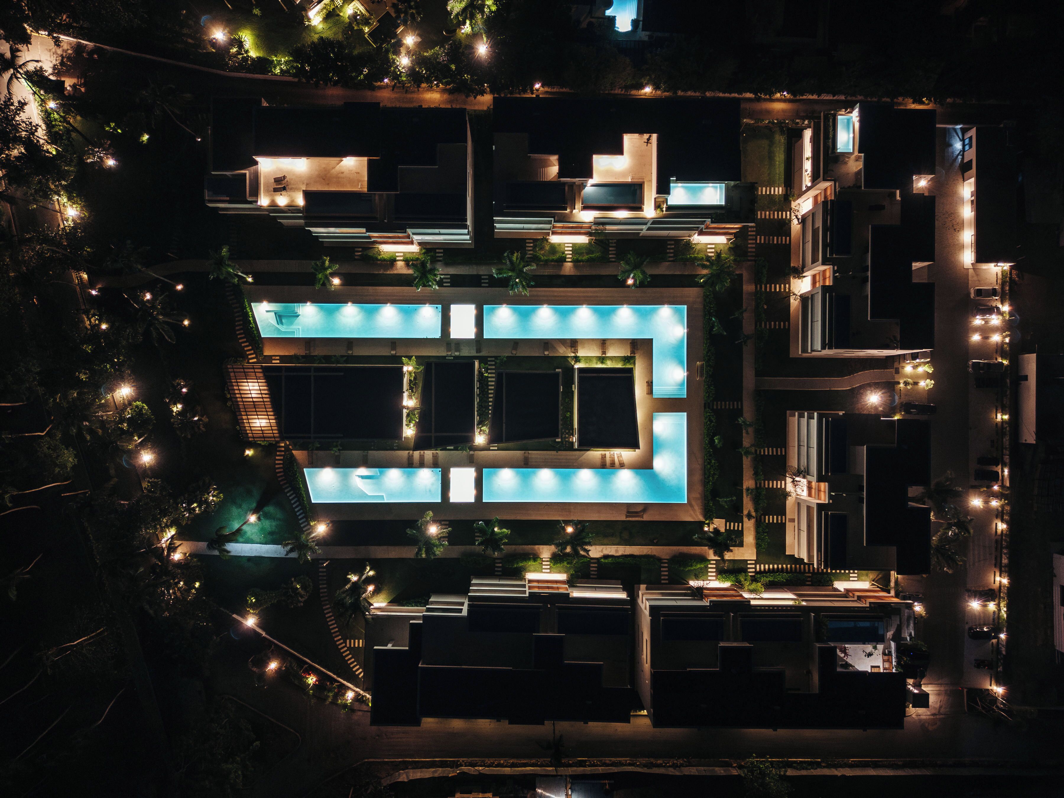 An aerial view of a modern hotel with a central swimming pool, multiple balconies, and surrounding greenery.