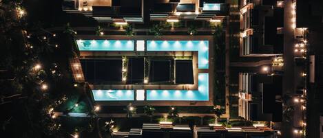 An aerial view of a modern hotel with a central swimming pool, multiple balconies, and surrounding greenery.