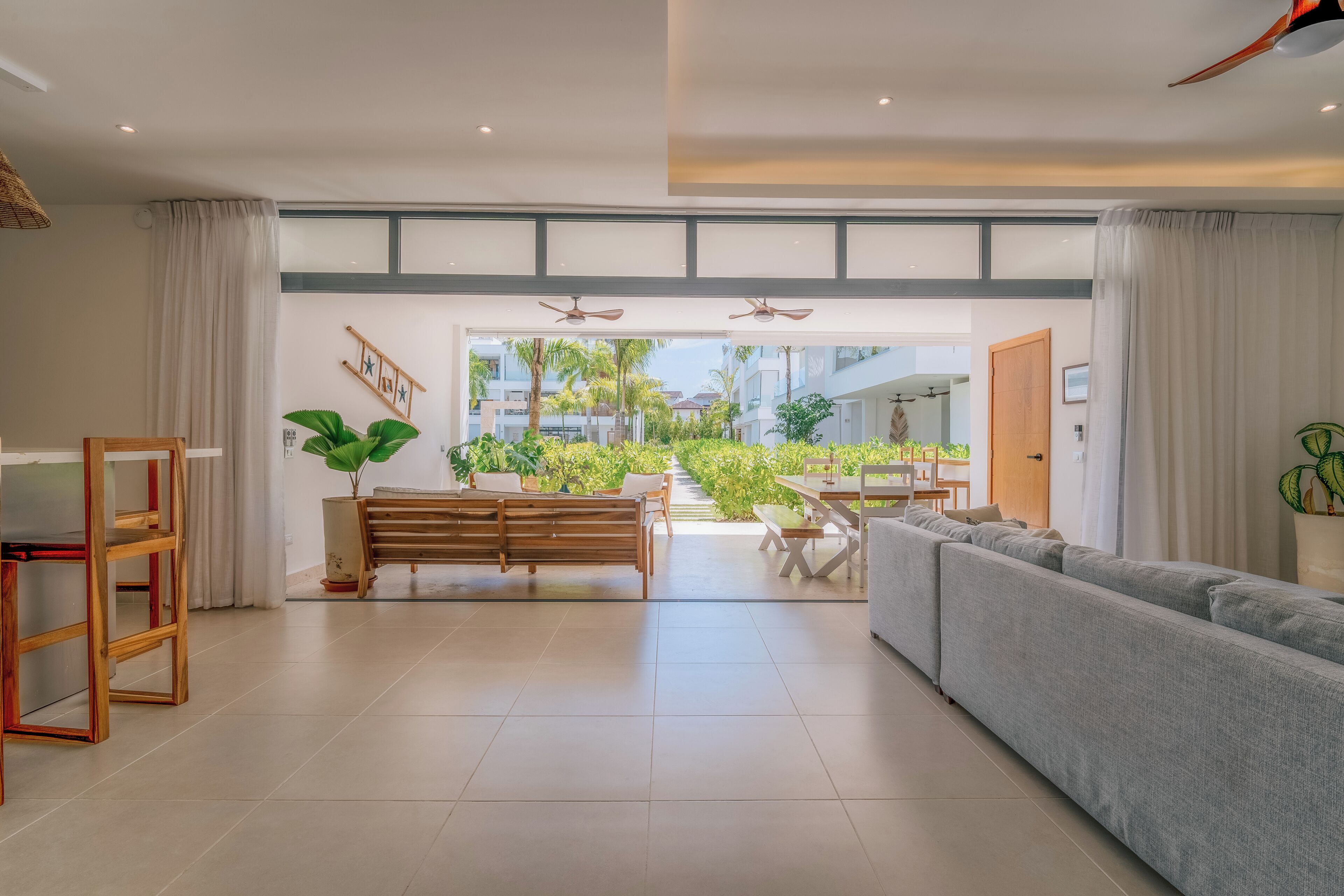 Amplia sala de estar con sofá, banco de madera y mesa de comedor. Se ve un jardín a través de un ventanal.