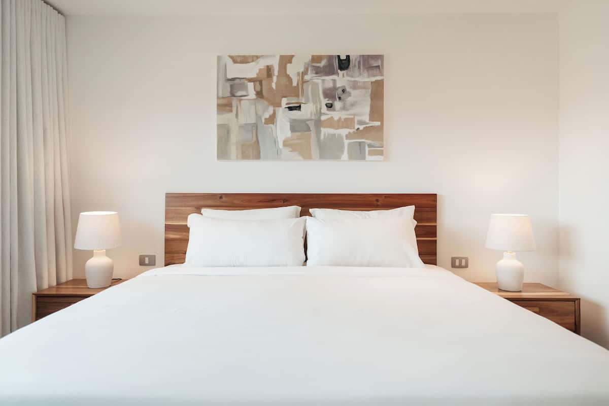 A hotel room with a large bed, two bedside tables with lamps, a wooden headboard, and a wall-mounted abstract painting.