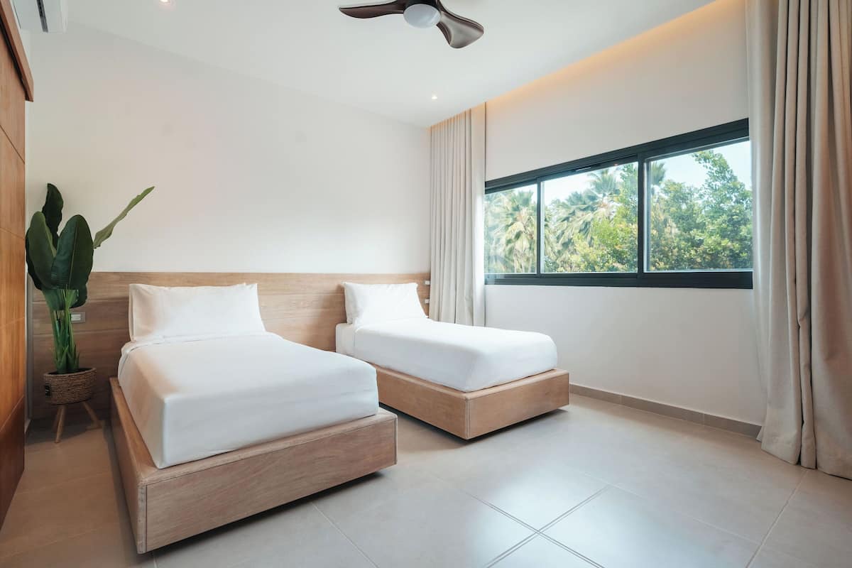 A modern bedroom with two beds, a ceiling fan, a large window with curtains, and a potted plant.