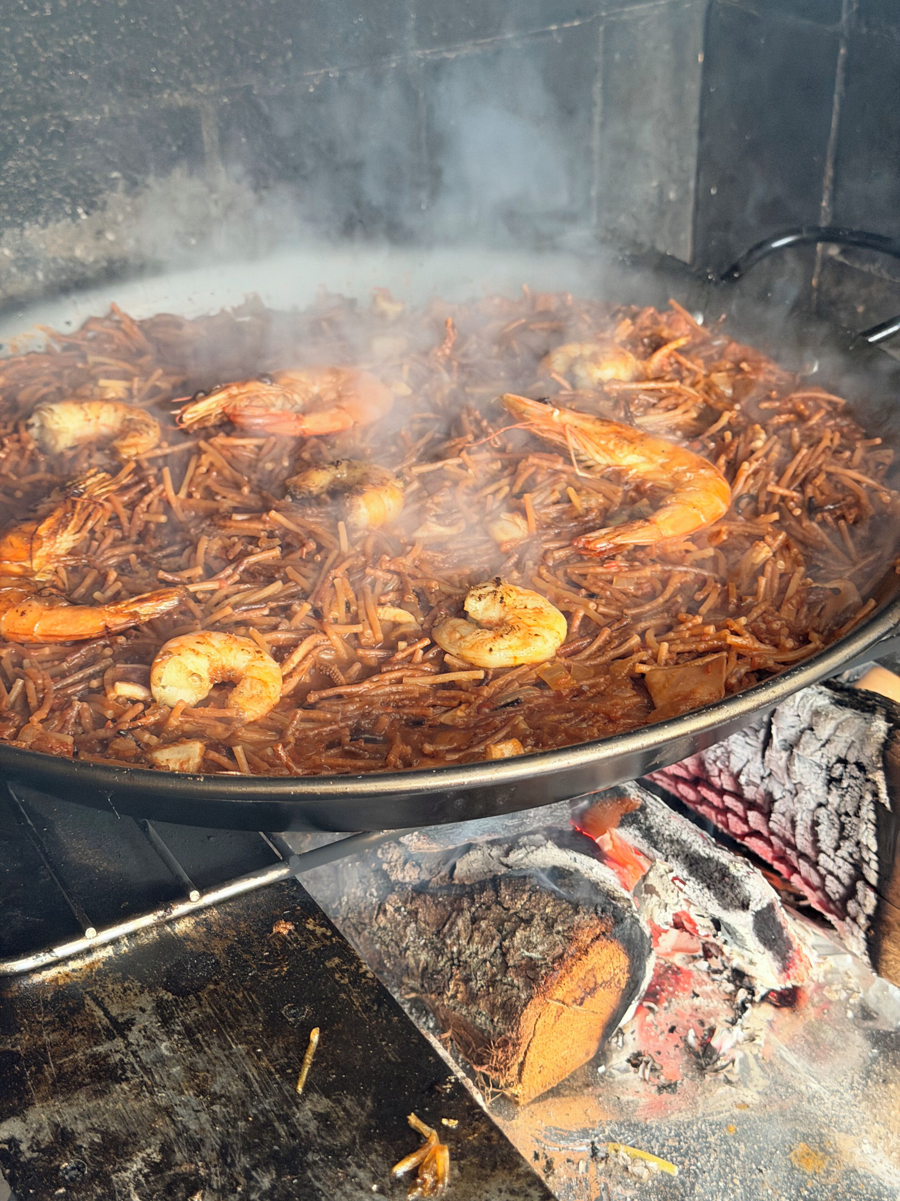 A large cooking pot filled with a dish containing shrimp and noodles, placed over an open flame.