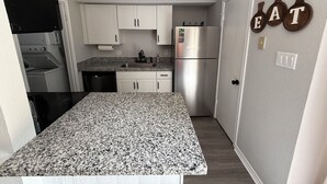 Eine kleine Küche mit weißen Schränken, einer Arbeitsplatte aus Granit, einem Edelstahlkühlschrank und einem an der Wand befestigten Schild mit der Aufschrift 'EAT'.