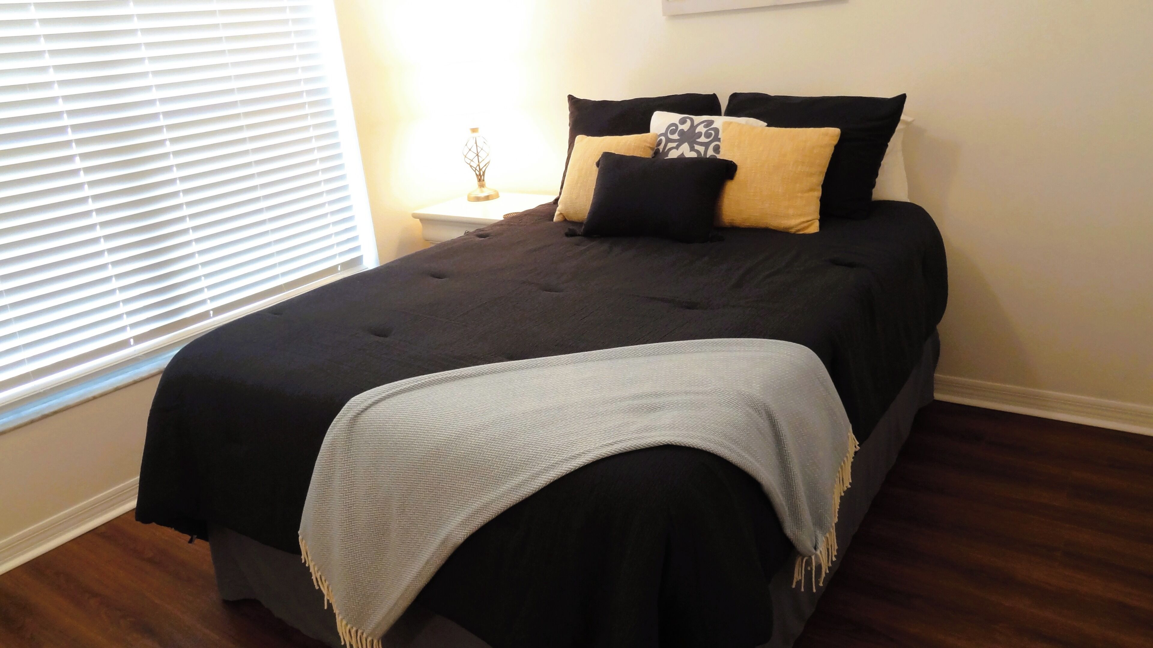 A bed with a black cover, a gray throw blanket, and multiple pillows in a room with wooden flooring and a window with blinds.