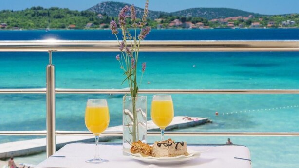 A table with two glasses of orange juice, a vase with flowers, and a plate with food, overlooking a turquoise sea and green hills. - Hotel Duje (Vodice)