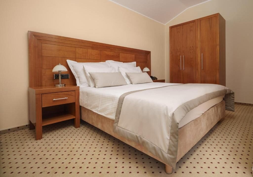 Quarto com cabeceira de madeira, cama com roupa de cama branca, mesa de cabeceira com abajur e guarda-roupa.