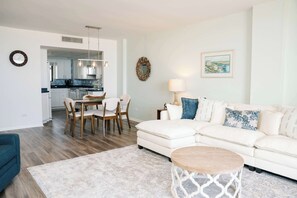 A modern living room with a white sofa, a dining table with chairs, a coffee table, and a kitchen in the background.