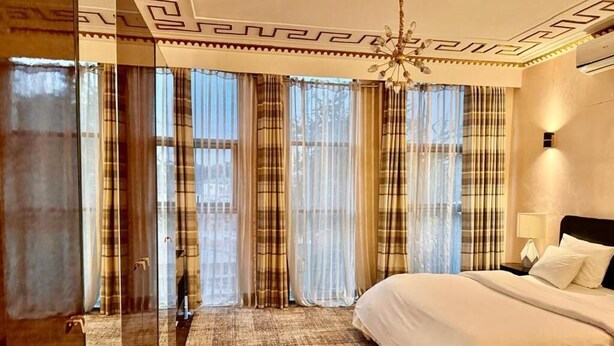 Een hotelkamer met een groot bed, een kroonluchter en uitzicht door dunne gordijnen.