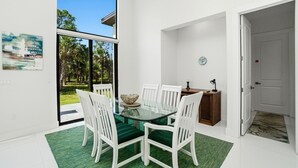 A dining area with a glass table, white chairs, a green rug, and a view of a garden through a glass door. - Christian Nature Haven (Naples)