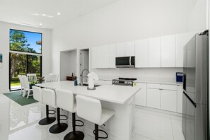 A modern kitchen with white cabinetry, a central island with bar stools, a microwave, and a refrigerator. - Christian Haven in Nature (Naples)