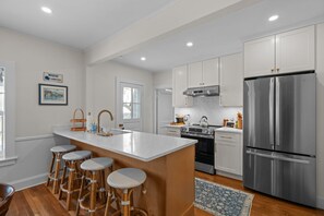 Eine moderne Küche mit weißen Schränken, einem Edelstahl-Kühlschrank, einer großen Kochinsel mit Sitzgelegenheiten und einem Essbereich mit Holzboden.