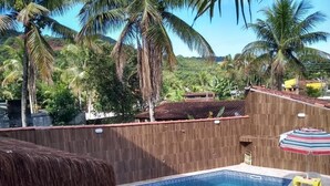 Une piscine dotée d’un toit de chaume et entourée d’une clôture en bois, bordée de palmiers et surplombant un paysage vallonné.