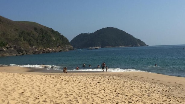 Une plage où l’on voit des gens se promener et nager, un ciel dégagé et une colline en arrière-plan.