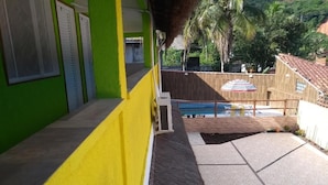 Un bâtiment à la façade verte et jaune, un espace piscine avec une terrasse carrelée, et une vue sur des palmiers et un bâtiment aux toits rouges en arrière-plan.