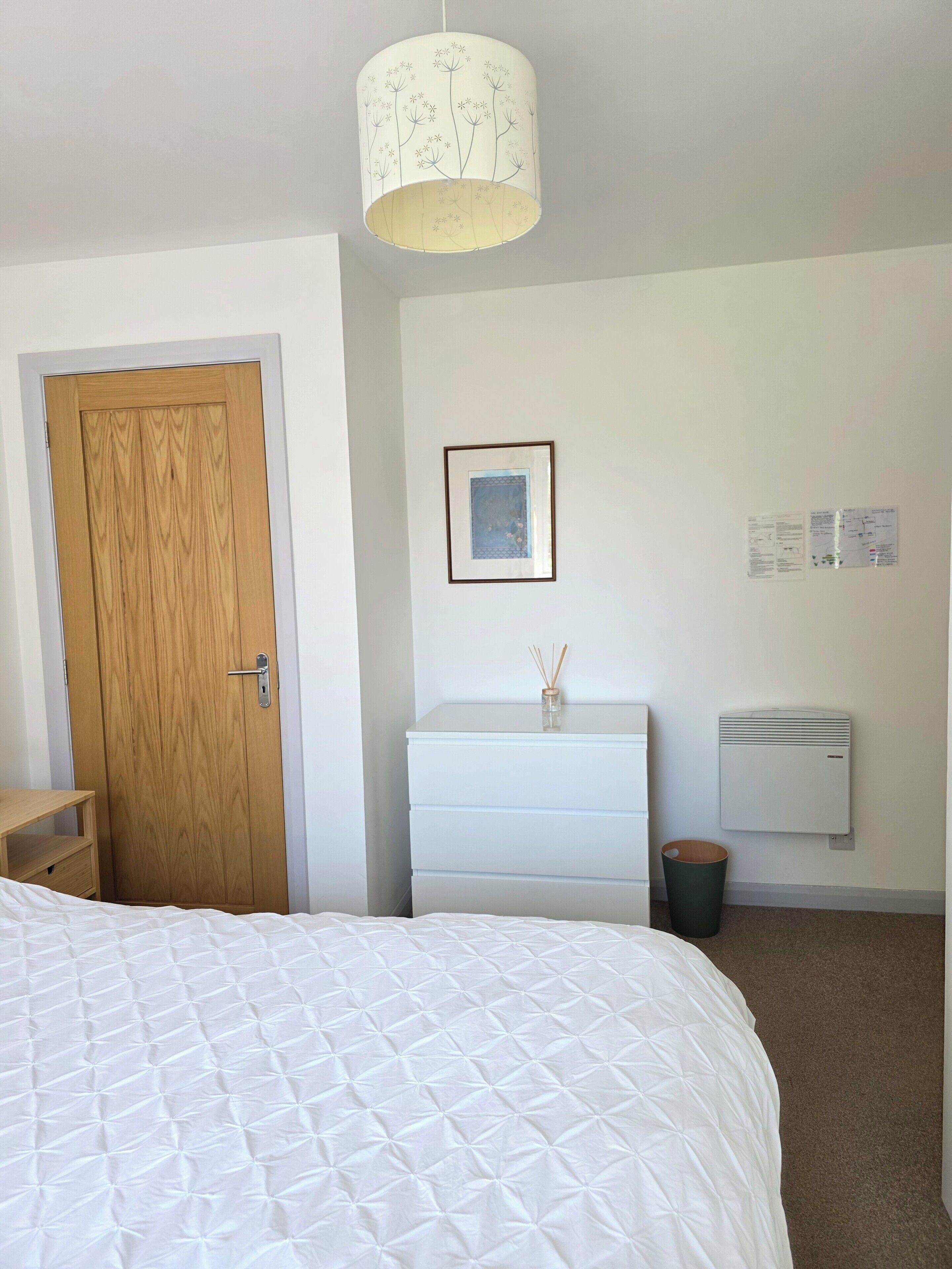 Une chambre à coucher avec un lit, une porte en bois, une commode blanche, un radiateur fixé au mur et un luminaire suspendu.