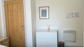 Une chambre à coucher avec un lit, une porte en bois, une commode blanche, un radiateur fixé au mur et un luminaire suspendu.