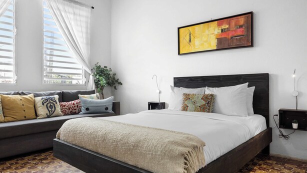 Un dormitorio con cama, sofá, planta, cuadro y piso con baldosas.