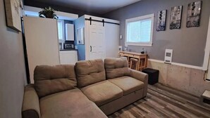 A living room with a beige sectional sofa, a small dining table, a kitchen area with cabinets and a microwave, and a sliding door closet. - Cozy Private Guest Suite Near SkyTrain (New Westminster)