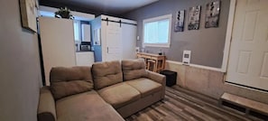 Un salon avec un canapé d’angle beige, une petite table à manger, un coin cuisine doté de placards et d’un four à micro-ondes, et un placard à portes coulissantes.