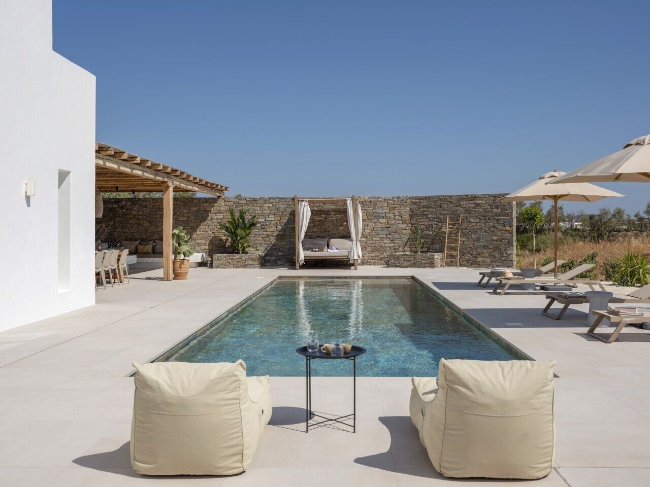 A pool area with lounge chairs, a table with drinks, and a stone wall in the background.