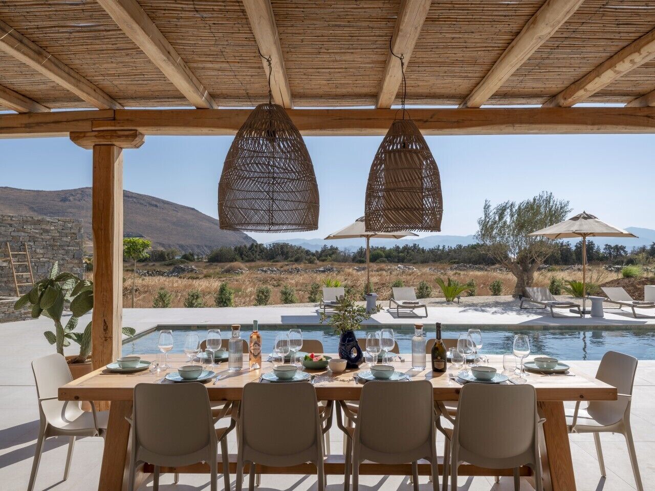 An outdoor dining area with a pool view, wooden pergola, and hanging light fixtures.