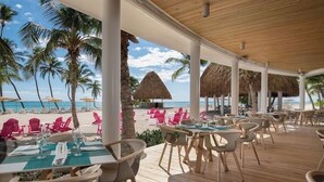 Een terras met tafels en stoelen, een houten terras en uitzicht op het strand en palmbomen.