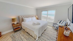 A bedroom with a large bed, a bedside table with a lamp, a chair, a TV, and a window with blinds. - Beautiful Destin on the Gulf 506 w/Beach Service (Destin)