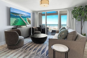 Una sala de estar moderna con un televisor grande, asientos cómodos, una mesa de centro redonda y vista al mar.