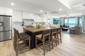 Una cocina moderna con armarios blancos, una isla grande con taburetes de bar, un refrigerador de acero inoxidable y vistas al océano a través de amplios ventanales.