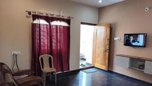 A living room with a sofa, a chair, a television, a wooden door, and a wall-mounted shelf. - Affordable Cozy Stay for Families Budget friendly (Kommaghatta)