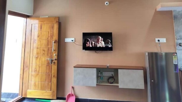 A room with a wooden door, a ceiling fan, recessed lighting, a mounted TV, a built-in shelf, and a refrigerator. - Affordable Cozy Stay for Families Budget friendly (Kommaghatta)