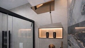 Uma casa de banho moderna com cabine de duche em vidro, parede de mármore, prateleira com champô e gel de banho, e torneira dourada.