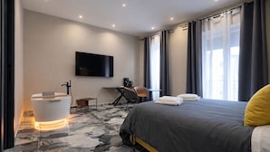 Quarto de hotel moderno com cama, escrivaninha, cadeira, televisão e banheiro com espelho e pia.