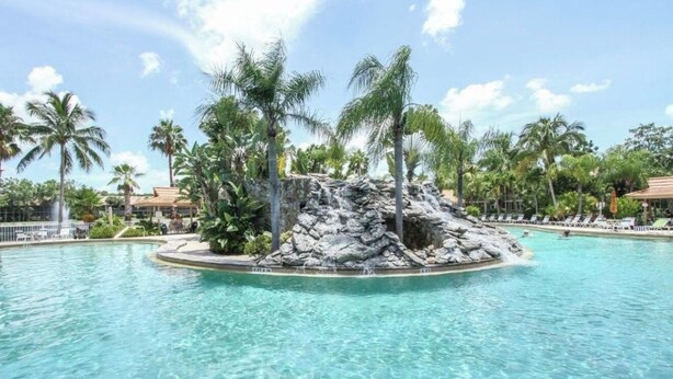 A pool with clear water, surrounded by palm trees and a rocky island feature. - Perfect Snowbird Retreat – Sunny Condo Near Pool & Clubhouse (Naples)