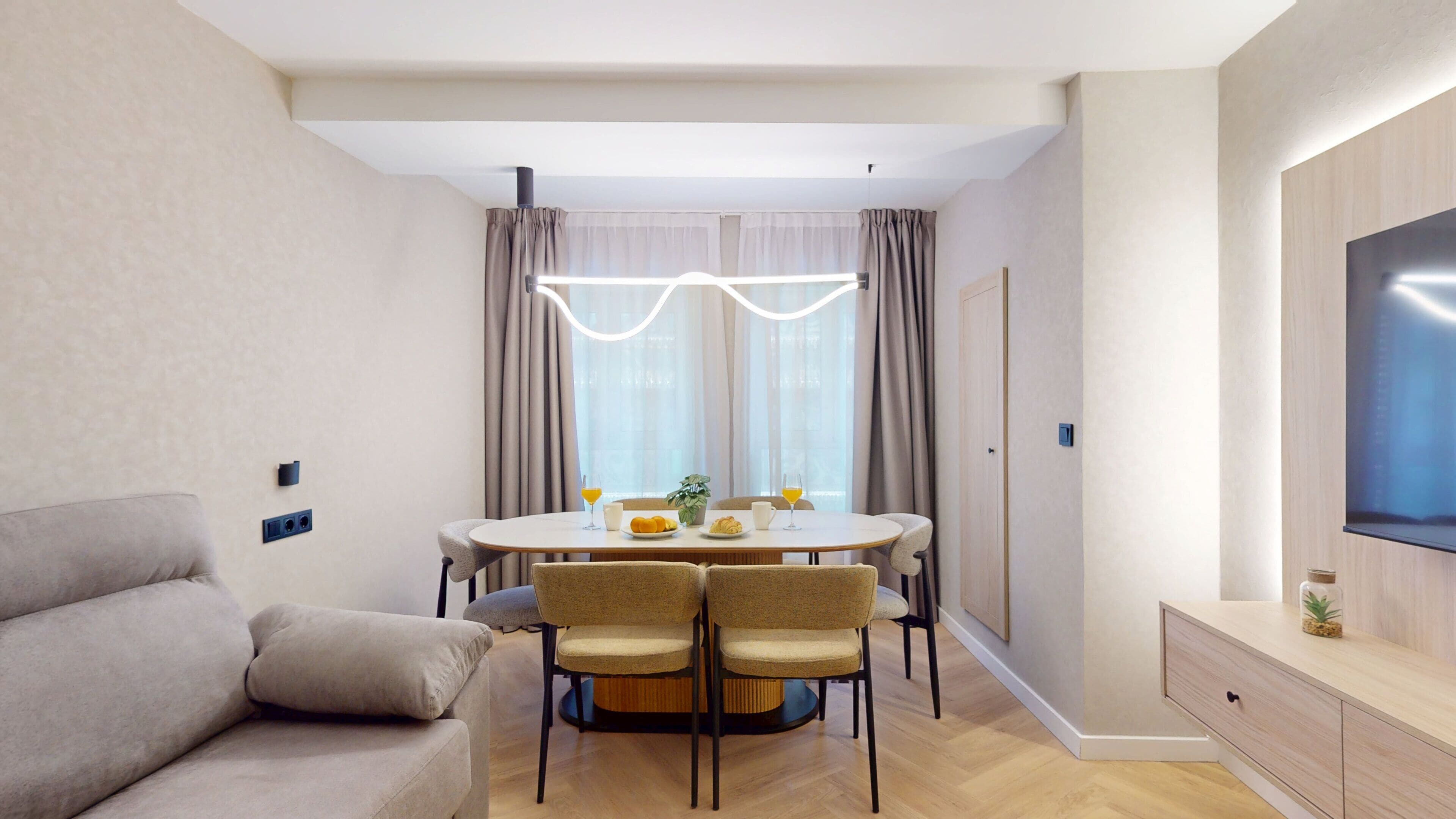 A modern living room with a sofa, a dining table with chairs, a flat-screen TV, and a large window with curtains.