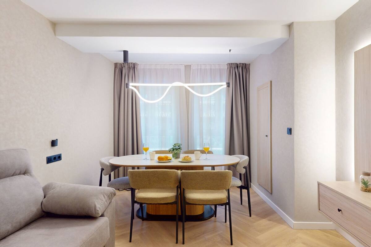 Una sala de estar moderna con un sofá, una mesa de comedor con sillas, un televisor y una ventana con cortinas.