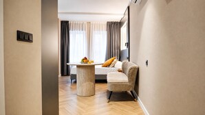 Een moderne hotelkamer met een bed, een rond tafel met fruit, een bank en uitzicht op het omliggende landschap.