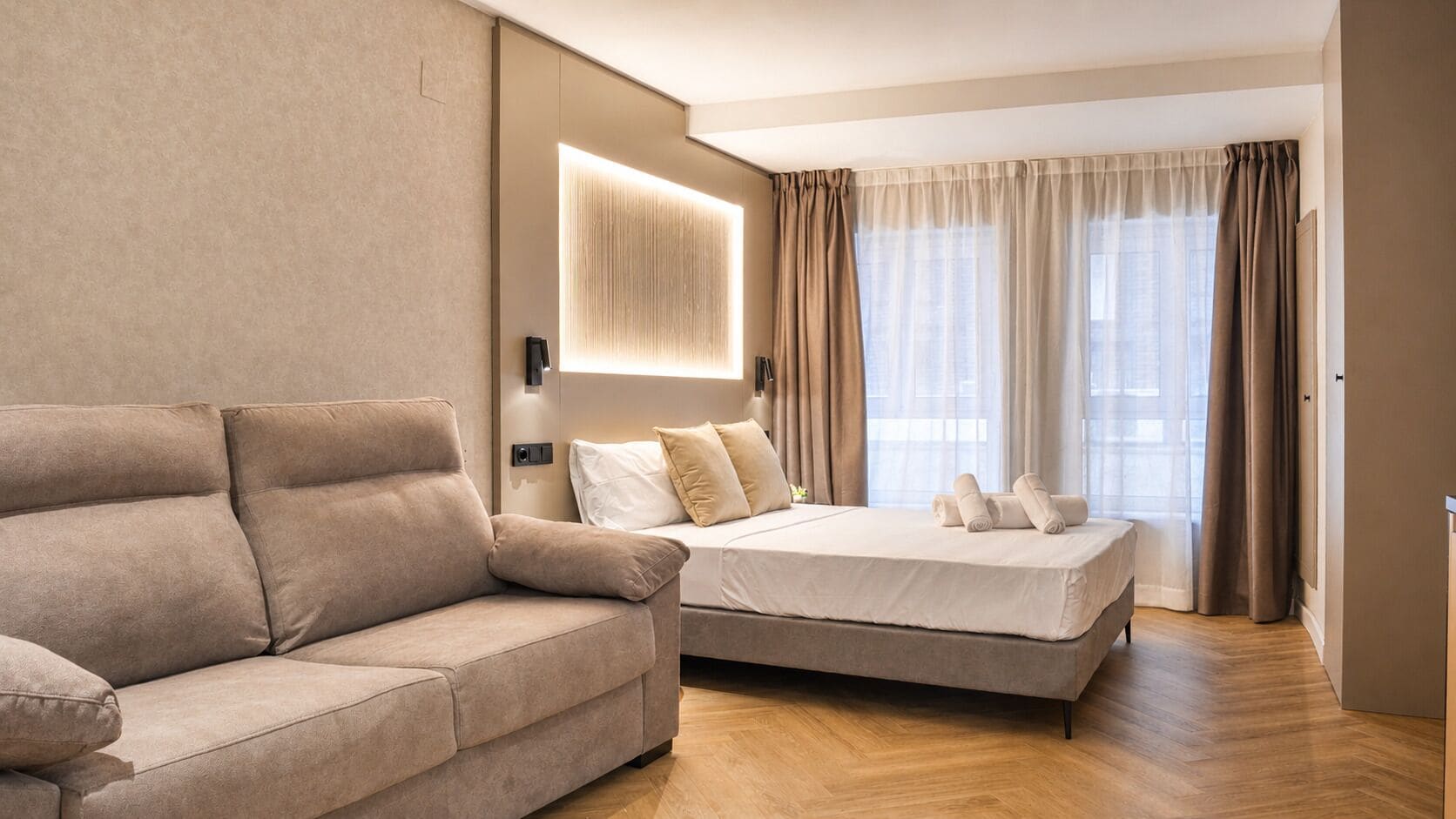 A modern hotel room with a sofa, a bed, a wall-mounted light, and a large window with curtains.