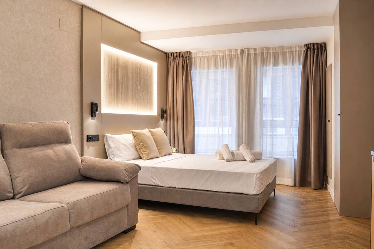 Una habitación de hotel moderna con sofá, cama, luz empotrada en la pared y ventanas amplias con cortinas.