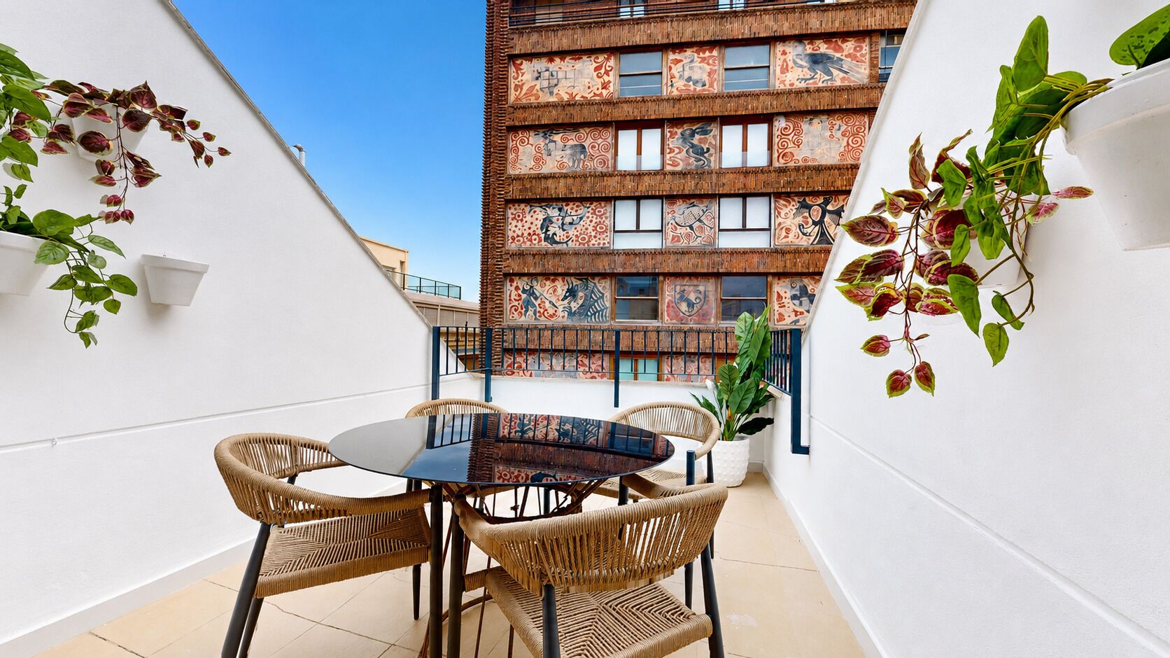 A balcony with a round table and wicker chairs, a potted plant, and a building with patterned windows in the background.