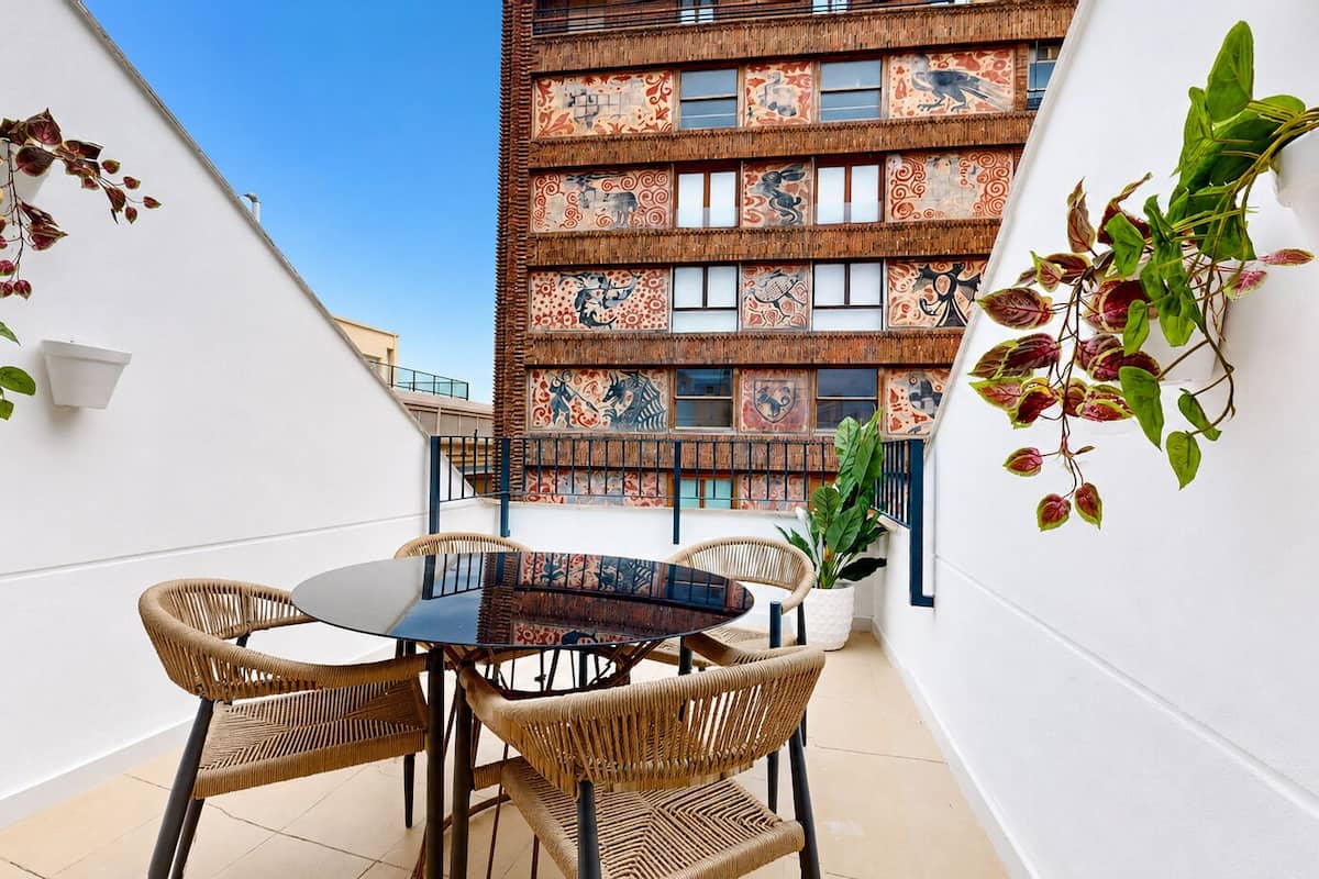 Un balcón con una mesa redonda y sillas de mimbre, una planta en maceta y un edificio con ventanas decoradas al fondo.