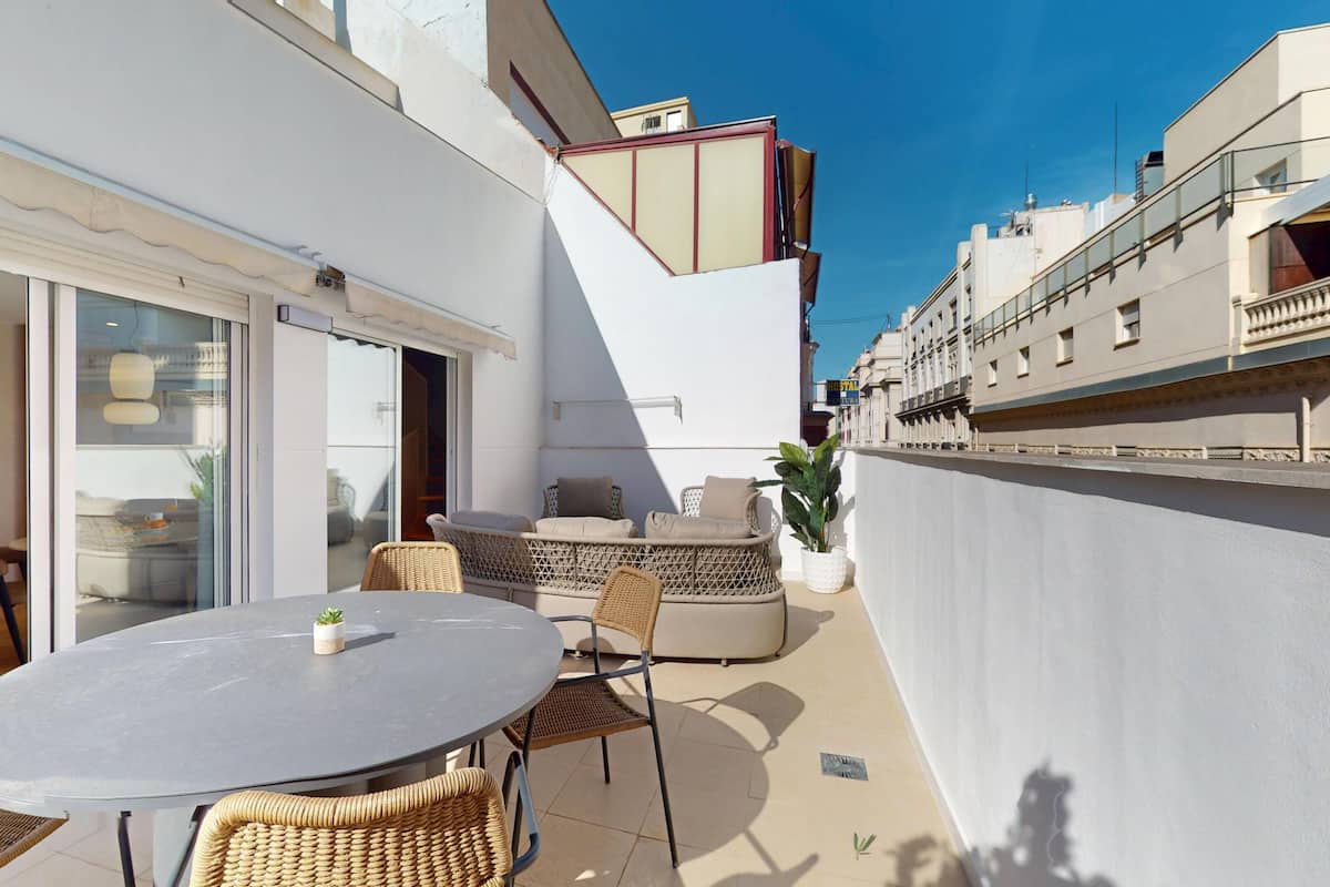 Un patio con una mesa redonda y sillas de mimbre, una puerta corrediza de vidrio que da a un área interior, y una pared blanca con balcón.
