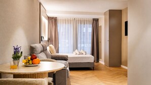 Een moderne hotelkamer met een bank, een bed, een eettafel met fruit en uitzicht op de stad door grote ramen.