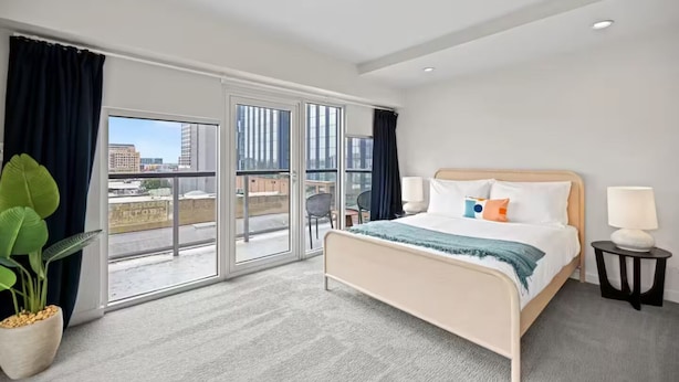 Habitación de hotel con cama, dos lámparas de noche, un ventanal con cortinas, una planta en maceta y vistas a la ciudad.