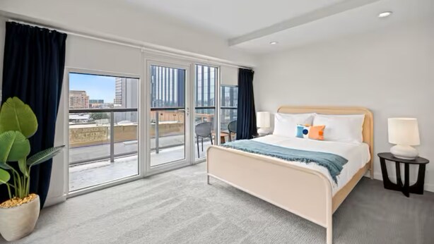 A hotel room with a bed, a nightstand, a potted plant, and a view of the city through large windows. - Perfectly Located - Cool 2 BR Apt - Downtown ATX (Austin)