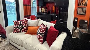 A living room with a sofa, a black ottoman, a ceiling fan, and a kitchen with black cabinets and a red countertop.
