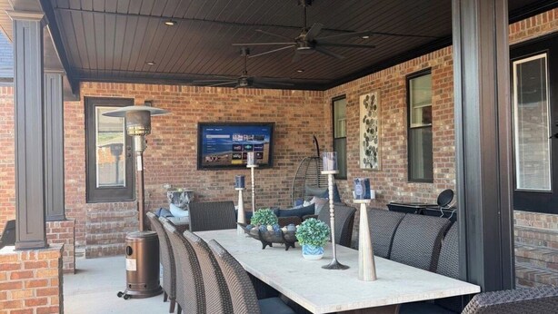 An outdoor patio with a brick wall, a flat-screen TV mounted on the wall, and a long table with wicker chairs.